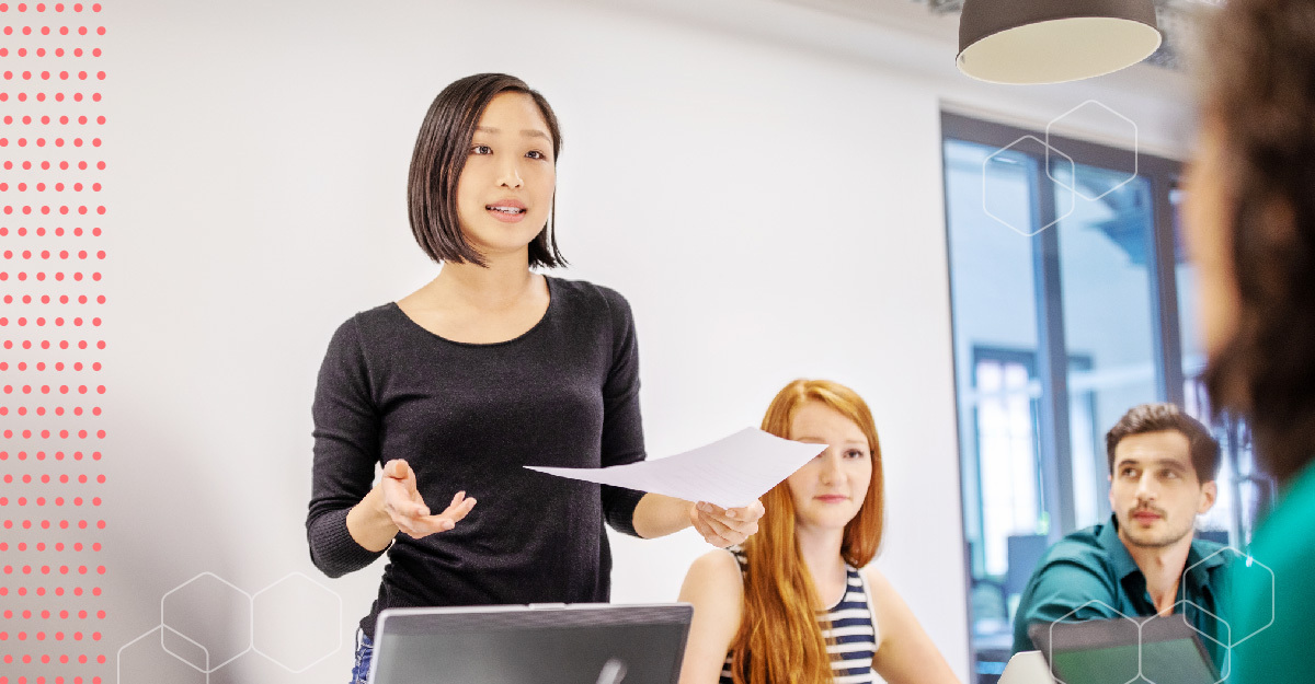 Woman standing at a table, giving a presentation to a group.