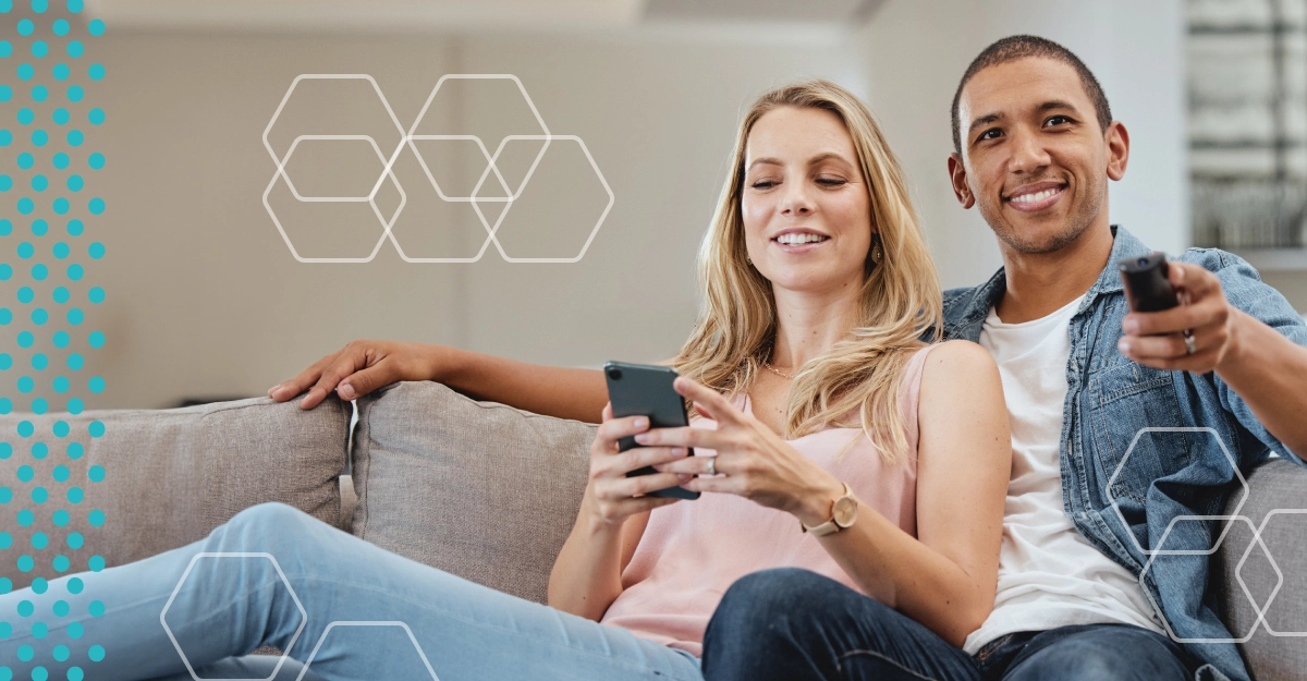 A woman looks at her smartphone and a man points his TV remote as they sit together on a couch